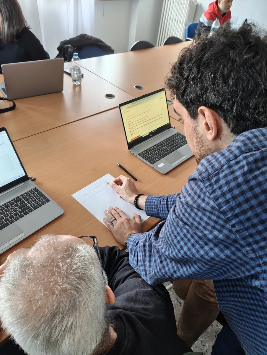 Il docente aiuta un partecipante seduto al tavolo, con carta e penna accanto al portatile