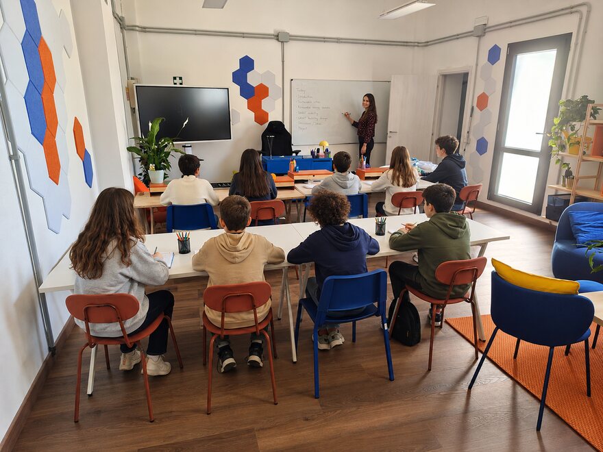 Illustrazione dell'aula durante una lezione: i partecipanti ai tavoli e la docente alla lavagna
