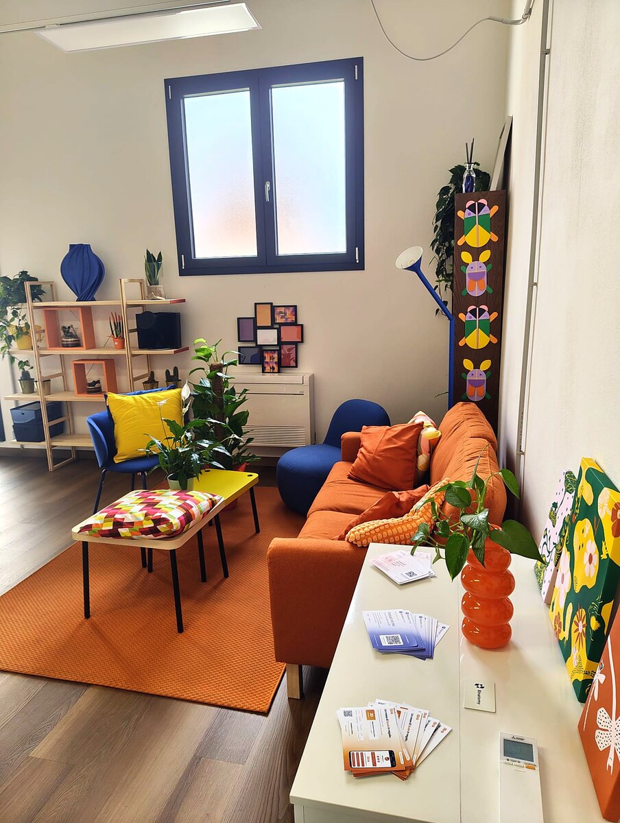 L'angolo lounge all'ingresso: divano arancione, libreria con le piante e il tavolo con i materiali